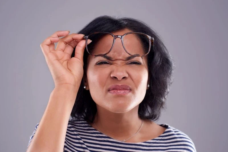 A woman squinting and holding a pair of glasses, representing the blurry distance vision symptomatic of nearsightedness.